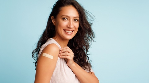 Symbolbild: Nach der Impfung zeigt eine Frau lächelnd auf ihr Pflaster auf dem Oberarm. © Shutterstock