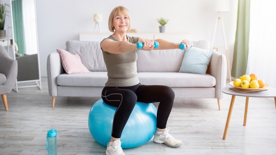 Eine Frau sitzt auf einem Fitnessball und hält Hanteln hoch. © Shutterstock